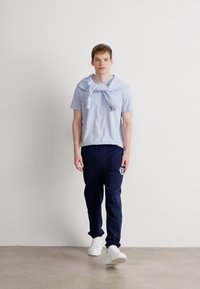 Világoskék, rövid ujjú póló, vállakon csomóval, sötétkék tréningnadrággal, amelyen logó található, és fehér sportcipővel.