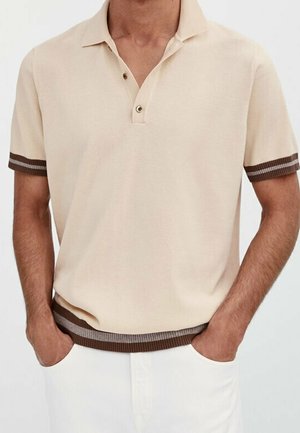 Beige Poloshirt mit kurzen Ärmeln, dunkelbraunen gestreiften Bündchen und Saum, getragen von einer Person mit den Händen in den Taschen einer weißen Hose.