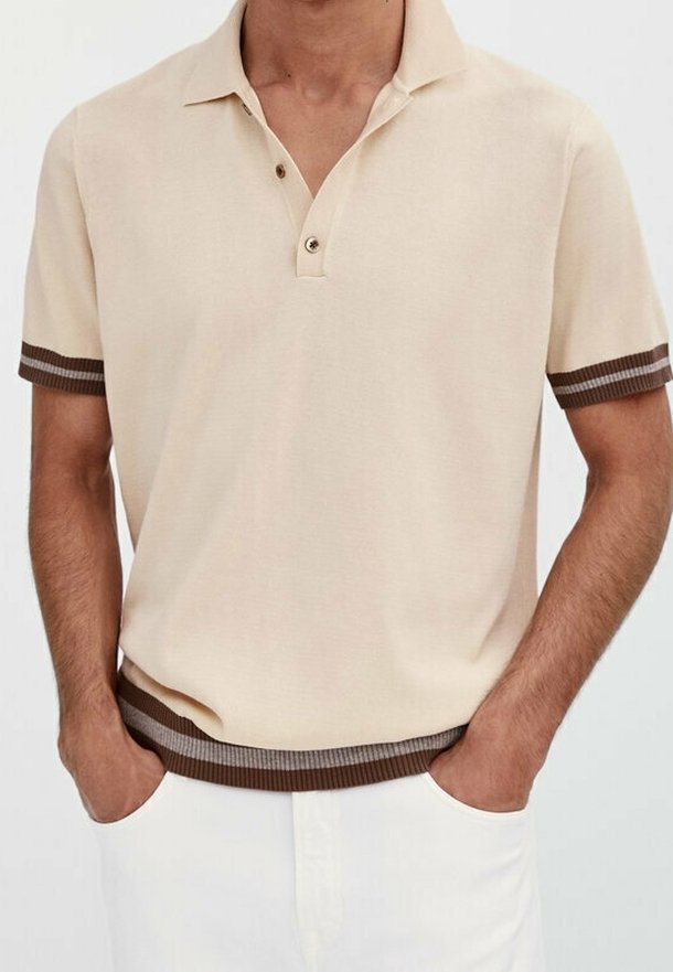 Polo beige à manches courtes avec poignets et ourlet rayés marron foncé, porté par une personne les mains dans les poches d'un pantalon blanc.