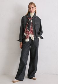 Donkergrijze spijkerjas met twee borstzakken en bijpassende wijde broek. Witte coltrui en rode sjaal met bloemenprint vormen een contrasterend accent.