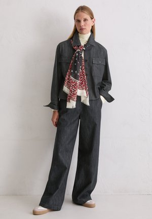 Donkergrijze spijkerjas met twee borstzakken en bijpassende wijde broek. Witte coltrui en rode sjaal met bloemenprint vormen een contrasterend accent.