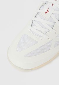 Scarpa sportiva bianca con tomaia in mesh, sovrapposizioni testurizzate e chiusura con lacci. Presenta un logo rosso e suola leggera in gomma.