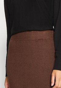 Gros plan sur une personne portant un haut noir à manches longues et une jupe brune côtelée à taille haute, avec sa main droite visible.