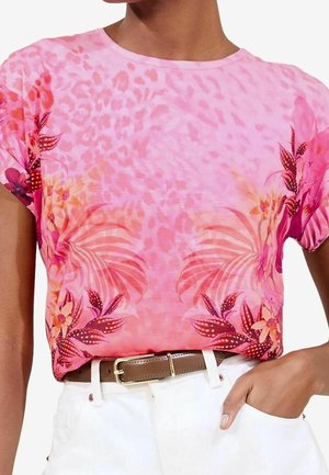 Persoon draagt een roze top met korte mouwen met bloemen- en dierenprint, gestopt in witte broek met een bruine riem, hand rust in de zak.