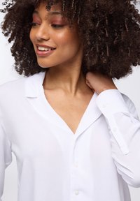 Camicia bianca con colletto a punta, maniche lunghe e bottoni in perla. Il tessuto leggero ha una consistenza liscia.