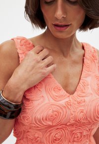 Coral sleeveless dress with textured rose patterns, featuring a V-neckline, complemented by glossy dark bangles on the wrist.