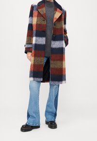 Cappotto in misto lana a quadri con strisce blu navy, rosse, gialle e bianche; design monopetto, ampio colletto e due bottoni. Abbinato a jeans a zampa d'elefante.