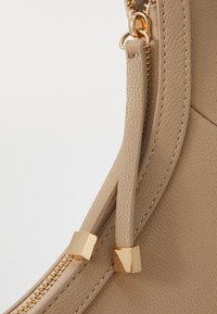 Sac à main en cuir beige avec une texture lisse, quincaillerie en métal doré et tirette de fermeture éclair discrète. Les coutures ajoutent de la dimension.