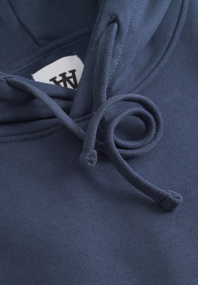Sweat à capuche bleu marine en tissu texturé. Il présente une fermeture à cordon de serrage rond et une étiquette avec un texte noir à l'intérieur de l'encolure.