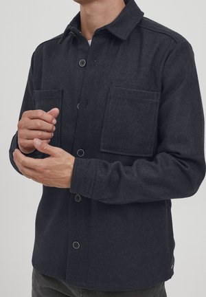 Homme portant une surchemise en laine gris foncé boutonnée avec deux poches poitrine, ajustant les manches sur un fond clair uni.