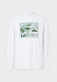 T-shirt bianco a maniche lunghe con un coccodrillo verde e il testo "LACOSTE," su uno sfondo a griglia. Realizzato in cotone, con una superficie liscia.