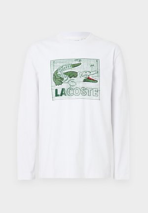 Witte lange mouwen T-shirt met een groene krokodil en de tekst "LACOSTE," op een achtergrond met een rasterpatroon. Gemaakt van katoen, met een gladde textuur.