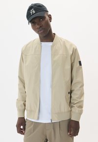 Chaqueta bomber beige con una textura suave, cierre de cremallera y bolsillos laterales. Combinada con una camisa blanca y una gorra negra con un logo.