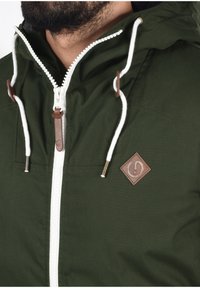 Giacca con cappuccio verde realizzata in tessuto, con zip bianca, dettagli in pelle marrone e cordini. Include un patch con il logo sul petto.