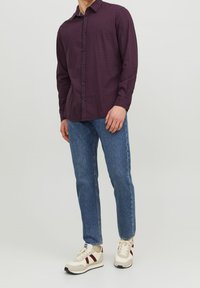 Mörkröd rutig långärmad skjorta, blå jeans och beige sneakers med röda accenter. Skjortan har knäppning framtill och spetsig krage.