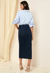 Lichtblauw poloshirt met opgerolde mouwen, gecombineerd met een donkerdenim kokerrok en sandalen met hoge hakken en bandjes. Achteraanzicht.