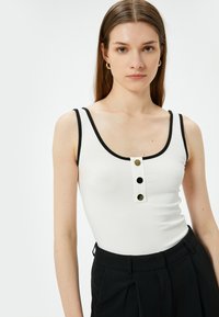 Hvid ribbet tanktop med sort kant, der har en scoop-udskæring og tre dekorative knapper foran. Glat tekstur og tætsiddende design.