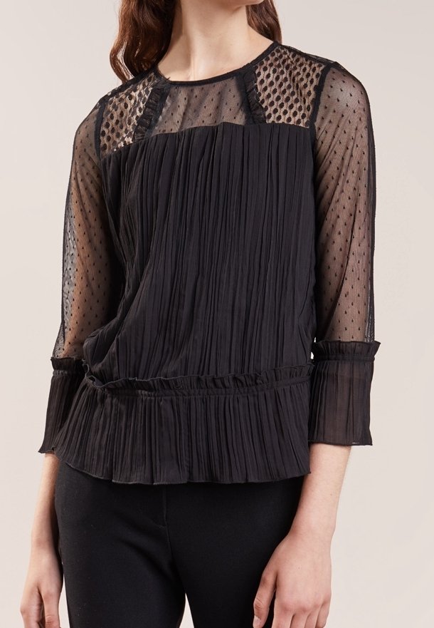 Femme portant un chemisier noir plissé avec manches et empiècement en tulle à pois, avec des détails à volants aux poignets et à la taille.