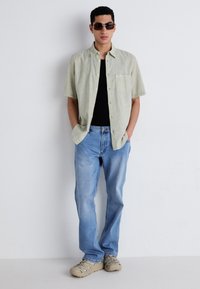 Chemise vert clair à manches courtes, t-shirt noir ajusté en dessous, jean bleu droit et baskets beiges, le tout accompagné de lunettes de soleil noires.