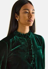 Samtgrüne Bluse mit rüschigem Stehkragen und Knopfleisten-Detail, aus glattem Material und mit langen Ärmeln.