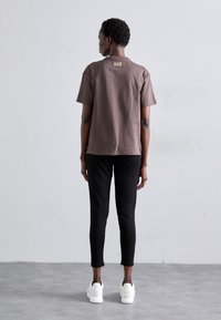 Person som står med ryggen mot kameran, iförd en brun kortärmad skjorta med logotyp, svarta byxor och vita sneakers mot en slät ljus vägg.