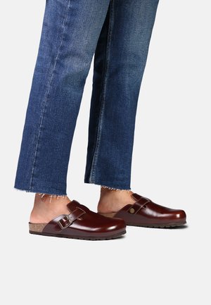 Person iført mørkeblå korte jeans og brune læder slip-on træsko med spænder, stående på en lysegrå overflade.