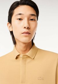 Pikétröja i beige, med en strukturert tyg, kort krage och två knappar, samt en tonad broderad logotyp på vänster bröst.