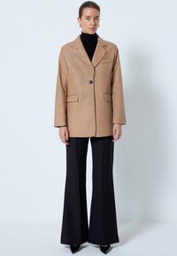 Beige læderblazer med ombukket krave, enkelt knaplukning og to forlommer, parret med sorte flagrende bukser og hæle.