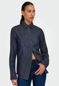 Tumman denimin nappikauluspaita, jossa on pitkät hihat ja hieman rento istuvuus. Sisältää etupaneelin ja ommeltuja yksityiskohtia.