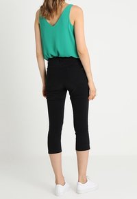 Žena s bezrukávovým zeleným topem, černými zkrácenými úzkými kalhotami a bílými teniskami, stojící zády k divákovi před jednobarevným pozadím.