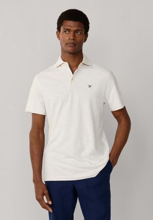 Hombre con una camisa polo blanca de manga corta con pequeño logotipo en el pecho y pantalones azul marino, de pie contra un fondo liso gris claro.