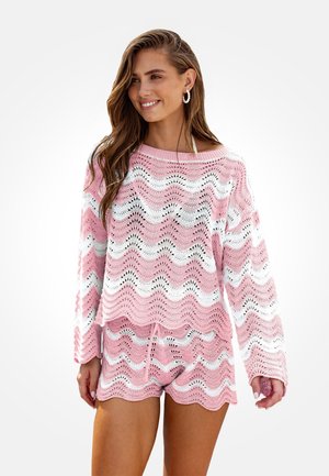 Femme souriante portant un pull en tricot à motif zigzag rose et blanc avec un short assorti, debout devant un fond clair uni.