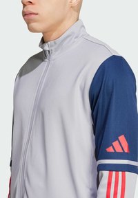 Chaqueta atlética gris con cuello alto, mangas azules y acentos rojos. El material es suave, con costuras visibles y logotipos.