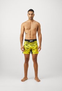 UFC márkájú sárga short, fekete derékpánttal, geometrikus mintázattal és sportos szabással, modell mezítláb egy semleges háttér előtt.