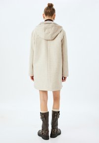 Abrigo con capucha de color beige claro hecho de una tela suave, con un corte relajado y mangas largas. Se lleva con botas de hebilla de color marrón oscuro.