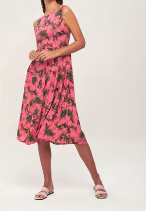 Donna che indossa un vestito midi floreale rosa senza maniche e sandali rosa chiaro, in piedi su uno sfondo chiaro e semplice.