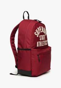 Mochila de tela roja con una parte superior redondeada, acentos negros, un bolsillo con cremallera en el frente y texto blanco en negrita "SUPERDRY STATE ATHLETICS" en la parte delantera.