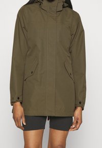 Person wearing an olive green hooded jacket with buttoned flap pockets and black shorts against a plain background.