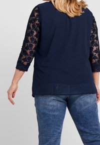 T-shirt navy à manches trois-quarts en dentelle avec un motif floral ; associé à un jean en denim délavé. Texture lisse et coupe décontractée.