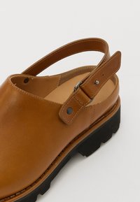 Scarpa slingback in pelle marrone con punta arrotondata, cinturino regolabile e suola in gomma nera testurizzata. Presenta cuciture minime e accenti in metallo.