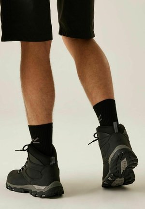 Osoba ubrana w czarne buty turystyczne, czarne skarpety i czarne szorty, stojąca na neutralnym tle, z jedną stopą lekko uniesioną.