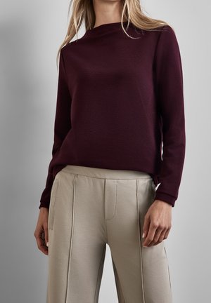 Kvinde iført en langærmet mørkelilla top og lys beige bukser med høj talje og sidelommer, stående mod en ensfarvet grå baggrund.