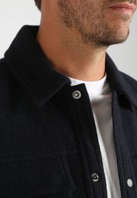 Close-up van de onderkant van het gezicht en de hals van een man die een donker wollen jasje draagt over een wit T-shirt met ronde hals.