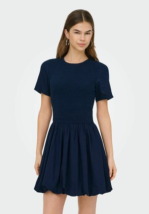 Femme aux longs cheveux bruns portant des boucles d'oreilles créoles et une robe à manches courtes bleu marine avec une jupe froncée, sur un fond clair uni.