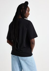 Black cotton t-shirt with short sleeves and a crew neck. The model wears light blue wide-leg jeans, showcasing the shirt's simple design.