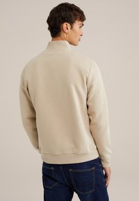 Felpa beige con colletto alto e polsini a costine. Il tessuto è morbido e ha una superficie liscia. Indossata con jeans blu scuro, conferisce un aspetto casual.