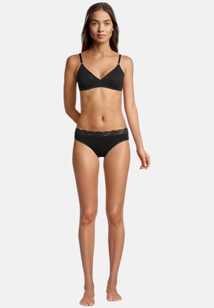 Woman standing barefoot wearing black wireless bra and black lace-trimmed underwear against a plain white background.