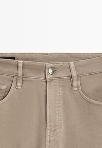 Gros plan sur la ceinture d’un pantalon en denim beige avec fermeture par bouton, passants de ceinture et coutures visibles de la poche avant.