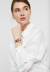 Montre en or rose avec un bracelet en maille, cadran rond et marqueurs d'heure minimalistes, portée au poignet d'une femme, contre une chemise blanche.