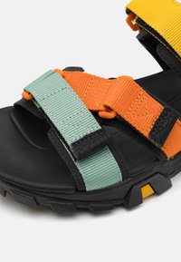 Sandalia negra con suela de goma texturada, que cuenta con tiras verdes, naranjas y amarillas de tela duradera. Incluye detalle de hebilla ajustable.